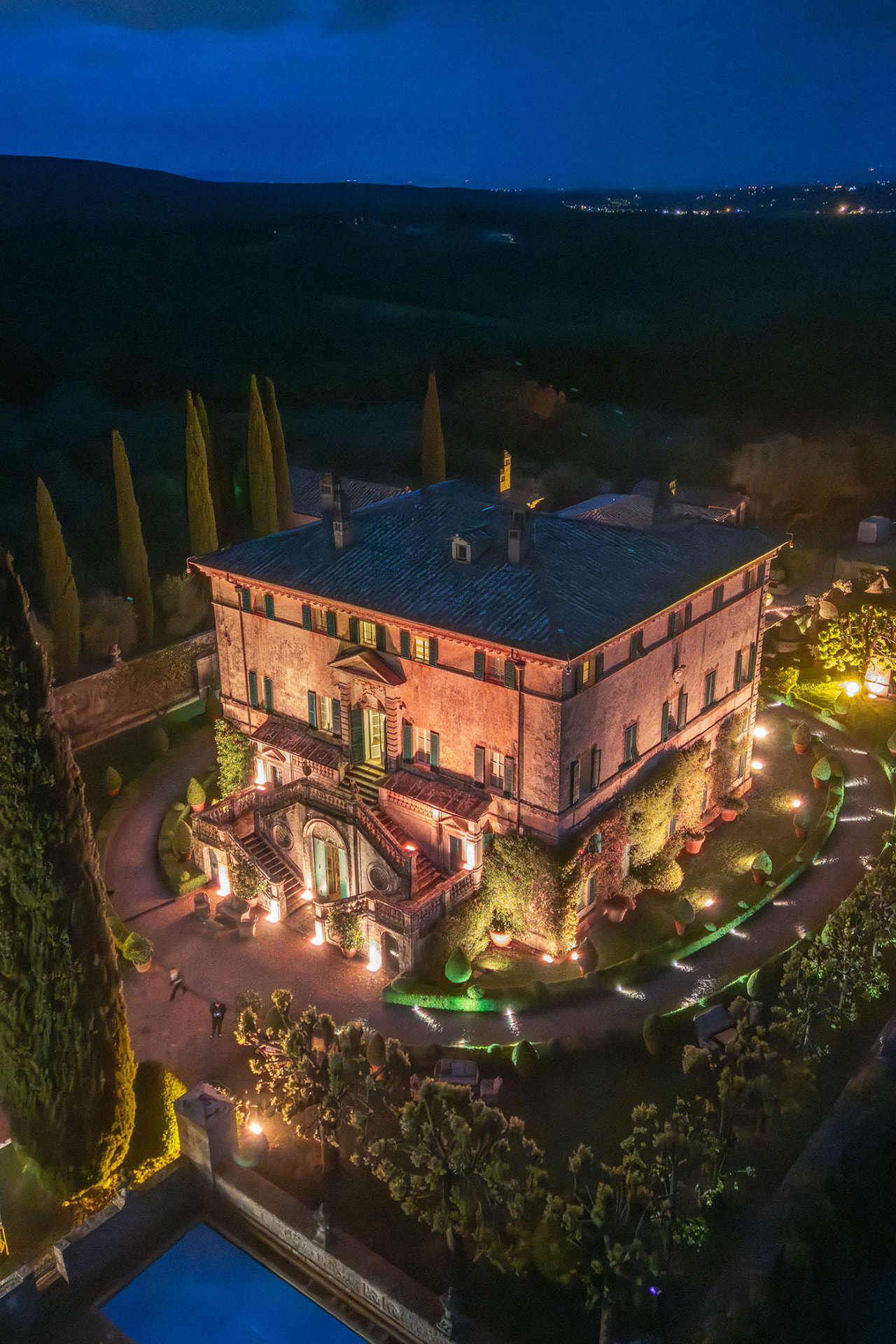 Aerial view of a candlelit Tuscan villa at dusk, cypress trees and clipped gardens around it